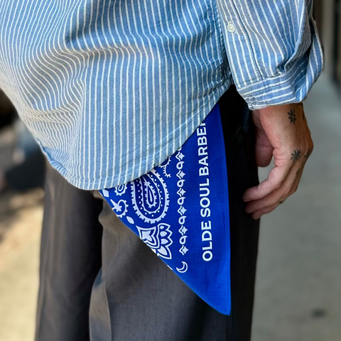 A person outdoors wearing a light blue and white vertically striped button-up shirt, dark gray trousers, and a bright blue bandana tucked into the back of the shirt. The bandana has white paisley patterns and the text 'OLDE SOUL BARBER'.