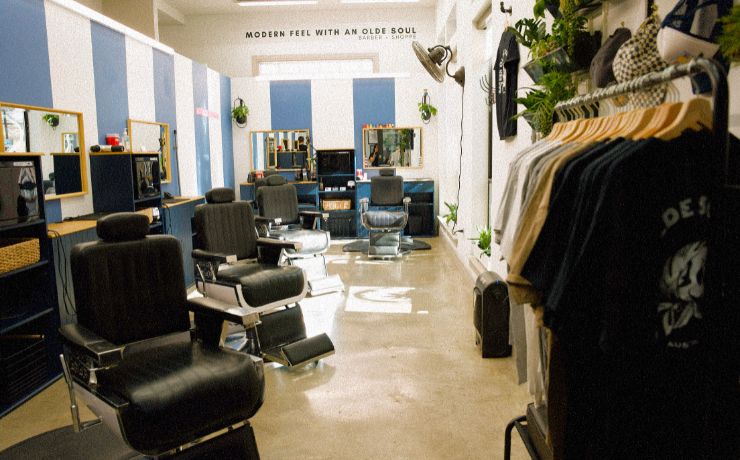 Interior of Olde Soul Barbershop East Six in Austin, TX, featuring modern black barber chairs, polished concrete floors, and sleek blue-and-white walls. A retail display with Olde Soul merchandise highlights the shop's stylish design, offering premium haircuts and grooming services in East Austin.